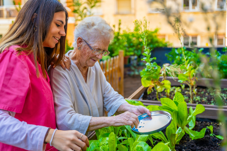 Caregiver helping a senior woman water plants in a raised garden bed