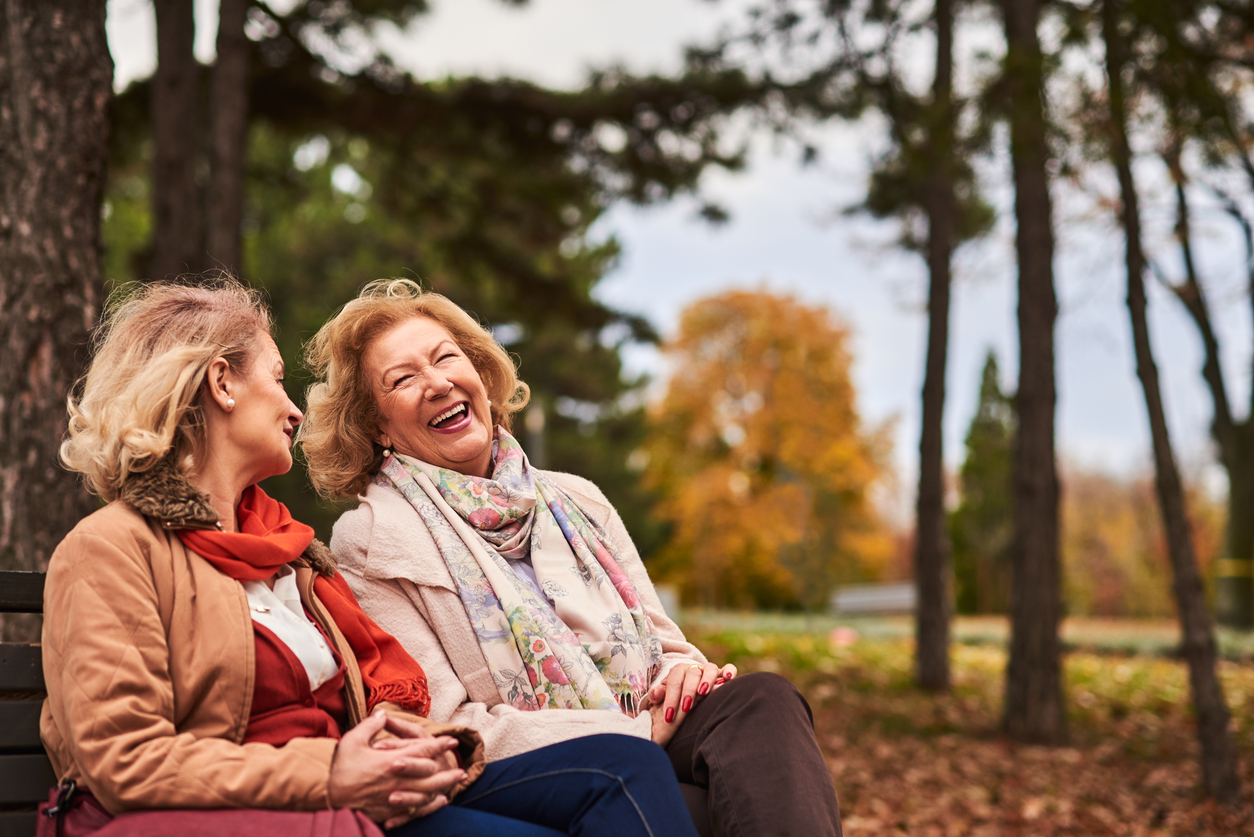 Two senior women laughing together on a park bench in autumn